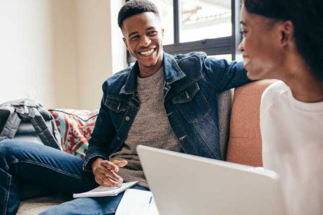 A young man is sitting on a sofa with a notepad smiling at a woman with a laptop.